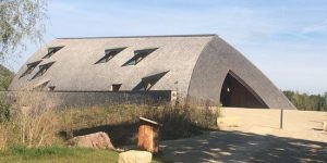 Curved brick building with a large arched entrance and small triangular windows, set in a sunny park.
